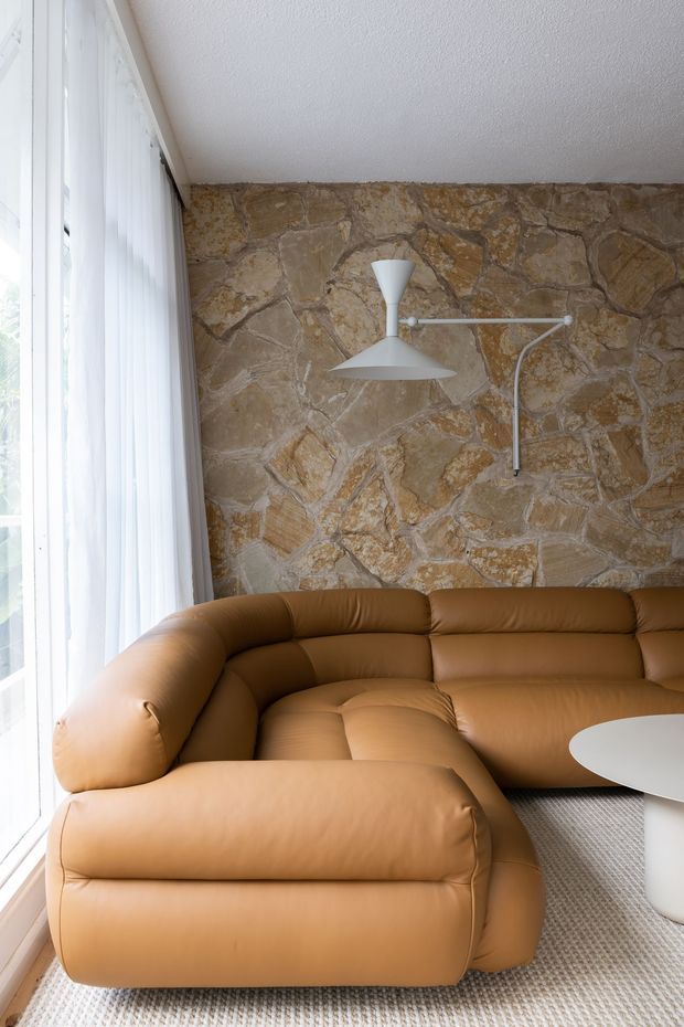 A bright living room corner with a large caramel-brown leather sectional sofa, white round coffee table, textured light carpet, full-height sheer window coverings, and a prominent irregular natural-stone feature wall. A white articulated wall lamp provides a sculptural modern accent.
