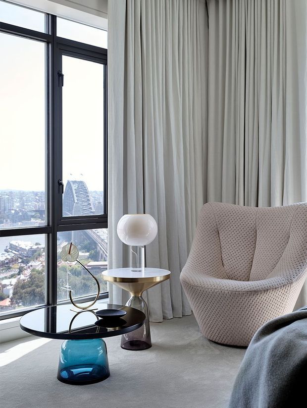 A refined high-rise interior corner with floor-to-ceiling black-framed glazing overlooking a dense cityscape. Full-height pleated ivory curtains frame a sculptural upholstered lounge chair, while a round black glass coffee table, translucent blue pedestal base, and elegant table lamp create a polished contemporary seating area. The room has a soft neutral palette with subtle metallic and jewel-tone accents.