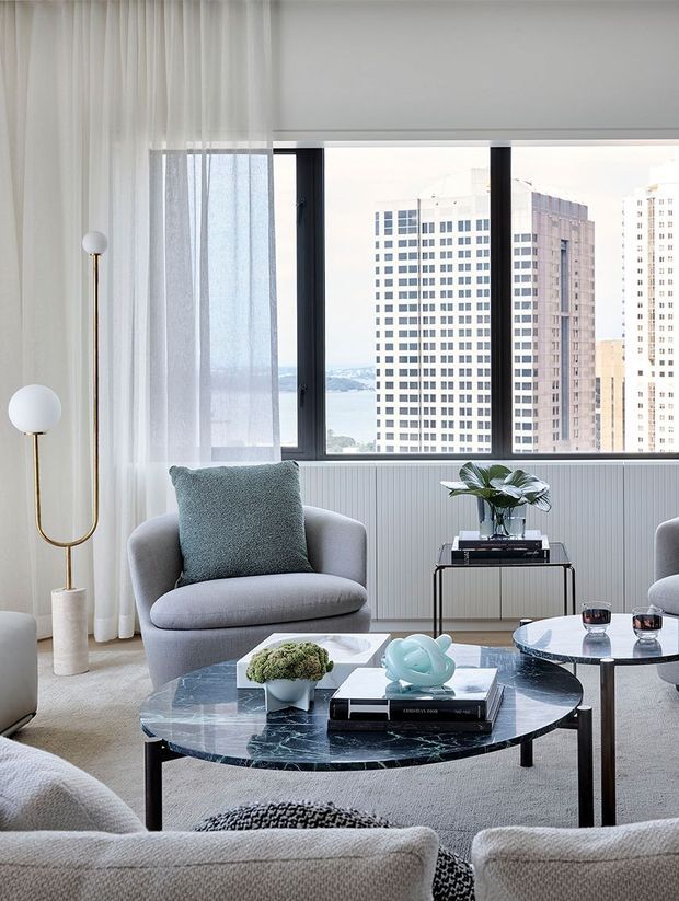 A refined contemporary living room with a large black-framed picture window overlooking a dense urban skyline. The room features softly upholstered pale grey seating, sheer white curtains, a dark marble oval coffee table, a sculptural globe floor lamp, a slim black side table, and restrained decorative objects in cool neutral tones.