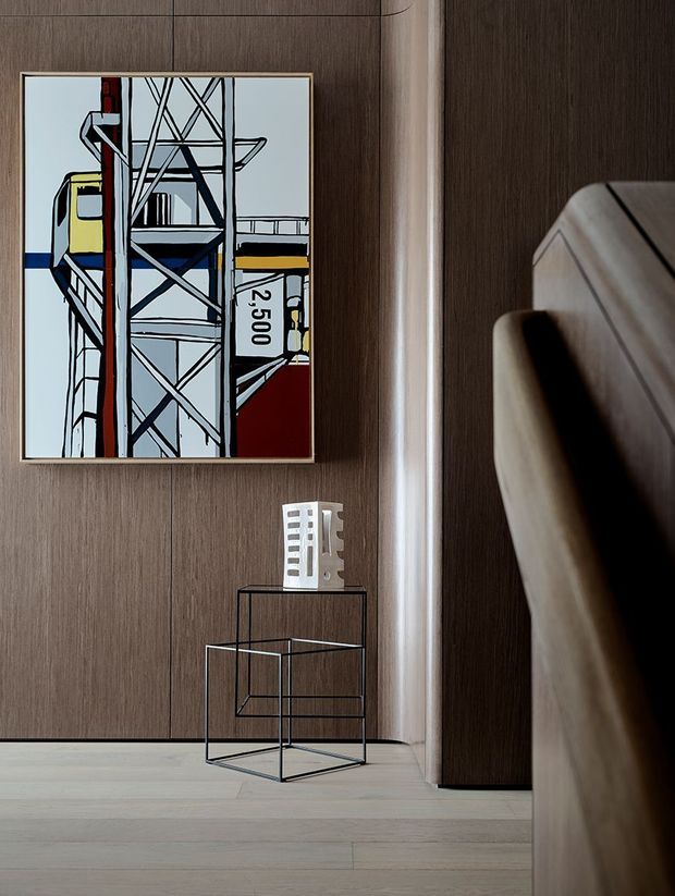 A refined minimalist interior vignette featuring a dark timber wall with vertical panel seams, pale timber flooring, and a slim black metal side table. A framed industrial artwork hangs above the table, while the right side is dominated by a curved upholstered or timber architectural element, likely part of adjacent furniture or built-in joinery. The palette is restrained in warm wood tones, black, white, and muted industrial colours.