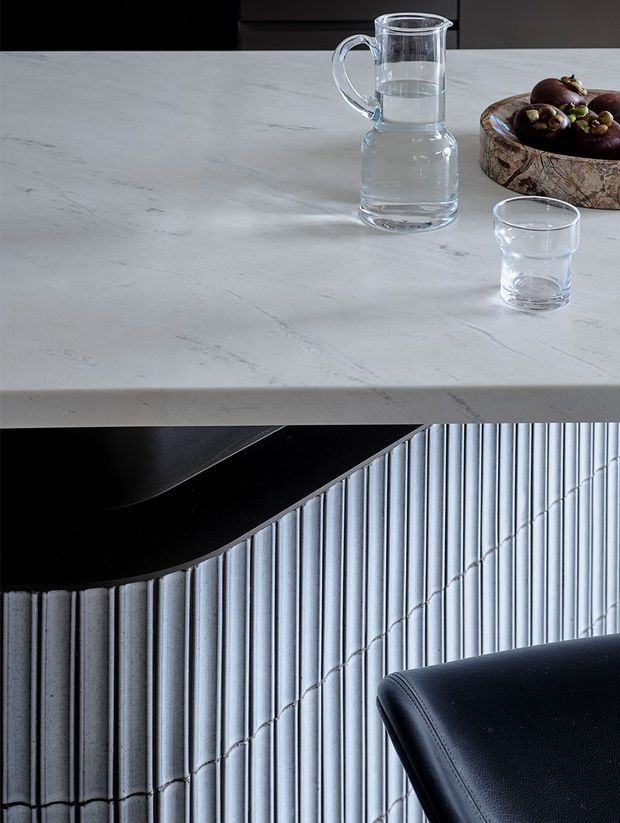 A split-detail image showing a pale grey-white veined stone benchtop with glassware and a wooden fruit bowl in the upper panel, and a close-up of vertically ribbed light-grey cabinetry or wall panelling with a dark curved benchtop and black upholstered seating in the lower panel.