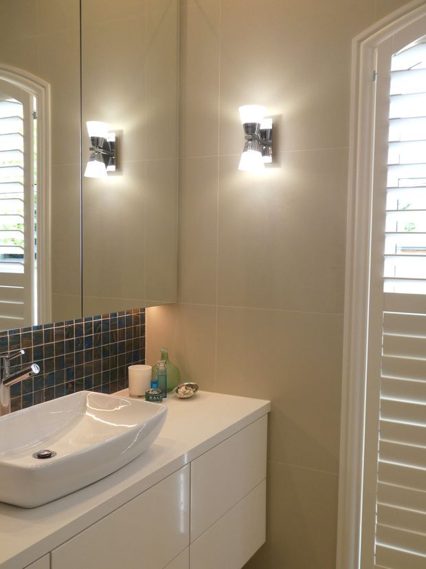 Bright contemporary bathroom vanity with a white vessel basin, white built-in drawers, large mirrored wall cabinet, beige tiled walls, and a dark blue-grey tiled splashback. Chrome wall sconces and white plantation shutters frame the space.