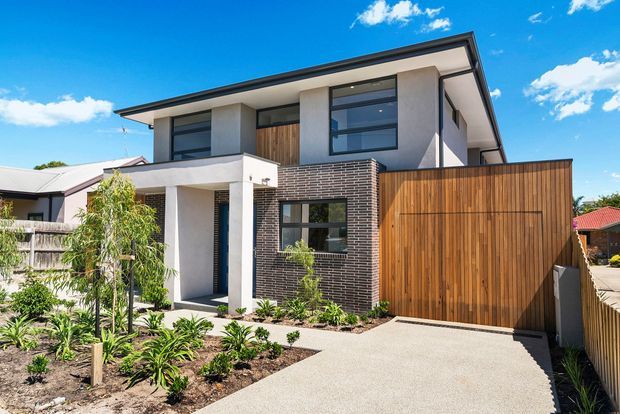 Contemporary two-storey residential house viewed from the street, featuring a low-pitched roof, white rendered walls, dark brickwork, extensive dark-framed glazing, timber-look upper cladding, and a large vertical timber garage door. A landscaped front garden and concrete driveway complete the exterior composition.