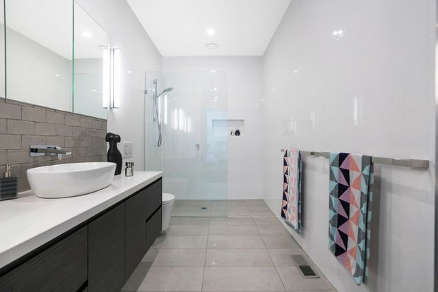 Bright contemporary bathroom with a long floating dark vanity, white vessel basin, large wall mirror, frameless glass walk-in shower, wall-hung toilet, and full-height glossy white wall tiles. Grey floor tiles and patterned towels add contrast.