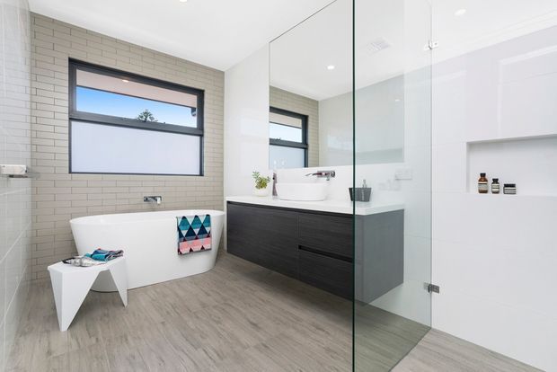 Bright contemporary bathroom featuring a freestanding white bathtub, wall-mounted dark timber vanity with a white vessel basin, large mirror, frameless glass shower enclosure, recessed shelving, light beige brick-look wall tiling, and timber-look flooring.