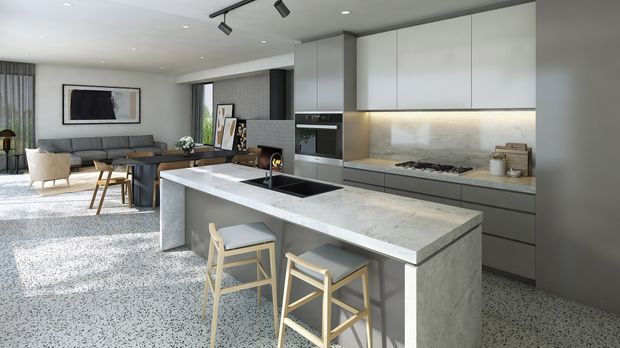 Contemporary open-plan kitchen and living area with a large pale stone island, integrated sink, two timber bar stools, flat-panel grey cabinetry, built-in appliances, and a stone-look splashback. A dining and lounge area with a sofa, armchairs, artwork, and large windows is visible beyond the kitchen. Track lighting and recessed ceiling lights contribute to the clean, modern fit-out.