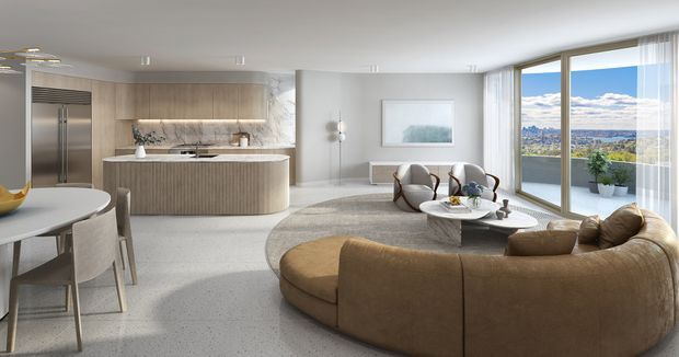 Open-plan contemporary apartment interior combining a light timber kitchen with a rounded island, dining area, and spacious living room. A large curved tan sectional sofa frames a low coffee table, while floor-to-ceiling glazing provides expansive city and water views. The palette is soft and neutral with warm timber, pale surfaces, and restrained modern furnishings.