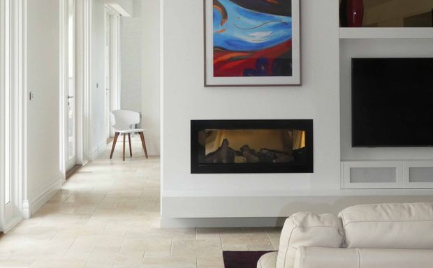 Bright contemporary living room with a white leather sofa, recessed gas fireplace, built-in media cabinetry and television, colourful abstract artwork, and pale stone or tile flooring. A glazed internal hallway or adjacent area with a small chair is visible to the left.