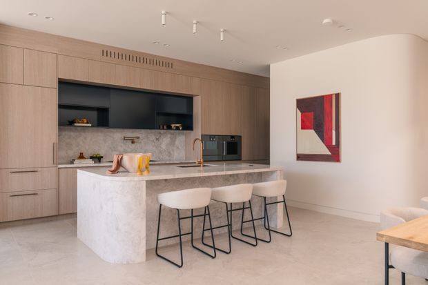 Contemporary open-plan kitchen with full-height light timber cabinetry, a dark recessed display and appliance wall, marble-look splashback and island, and three pale upholstered stools with black metal frames. The kitchen opens toward a partially visible dining or living area, with neutral plaster walls, integrated lighting, and a bold abstract artwork adding a red accent.