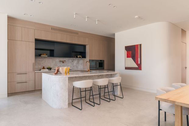 Contemporary open-plan kitchen and dining area with extensive light timber cabinetry, a large pale stone island with waterfall end, integrated dark shelving and appliances, and a marble-look splashback. Three bar stools line the island, while a timber dining table and upholstered chairs occupy the foreground. The room has a light neutral palette, recessed and track lighting, and minimal decorative detailing.