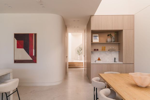 A refined open-plan dining and kitchenette area with a warm light-wood built-in cabinet wall, integrated shelving, pale stone benchtop, and a timber dining table with upholstered chairs. The room has smooth curved plaster walls, a light polished floor, recessed ceiling lights, and an abstract artwork. A short stair and bright hallway are visible beyond.