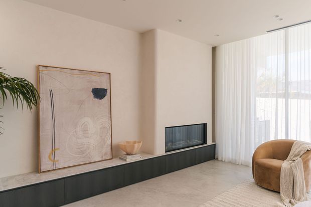 Bright contemporary living room with a long low dark built-in media cabinet and recessed fireplace, pale stone or concrete-look surfaces, neutral plaster walls, floor-to-ceiling sheer curtains, and a tan rounded armchair with a throw. A large abstract artwork and indoor palm add restrained decorative accents.