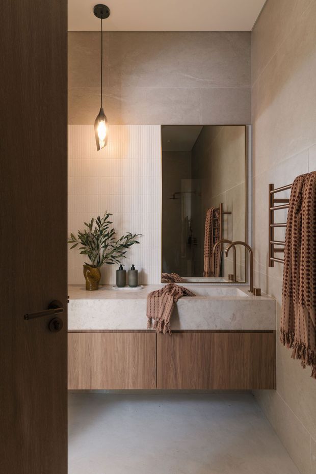 A warm contemporary bathroom viewed through a partially open dark timber door. The compact room features a floating timber vanity with a pale stone benchtop, integrated basin, brushed brass tapware, a large mirror, warm grey wall and floor tiles, a white vertically ribbed feature tile wall, pendant lighting, and a brass towel rail with a textured terracotta towel. A small decorative plant and accessories sit on the vanity.