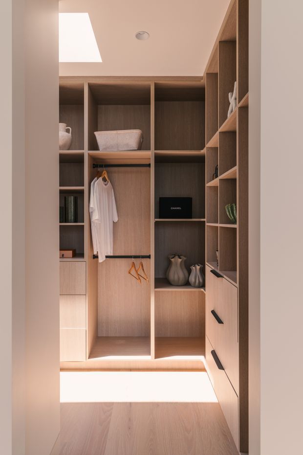 A compact walk-in wardrobe with floor-to-ceiling built-in timber-look shelving, open hanging sections, drawers, and display niches. Neutral pale wood joinery, a skylight, recessed ceiling light, and integrated lower lighting create a warm, refined dressing space.