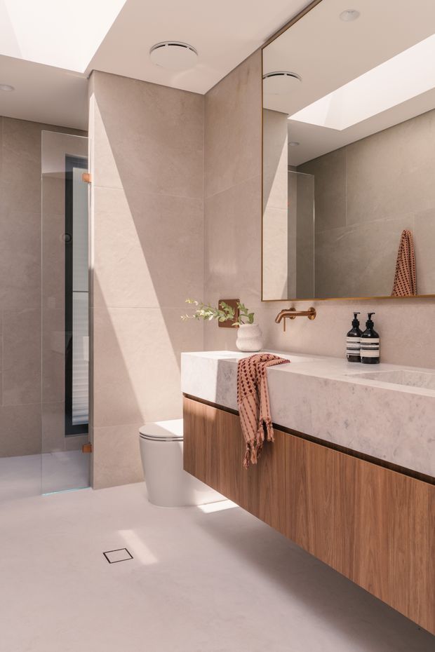 A contemporary bathroom featuring large beige stone-look wall and floor tiles, a frameless glass shower, wall-hung toilet, floating timber-veneer vanity, and a pale stone double basin benchtop. A large mirrored cabinet spans the vanity, with warm brass tapware and understated black accessories. A skylight or clerestory opening brings strong natural light into the refined space.