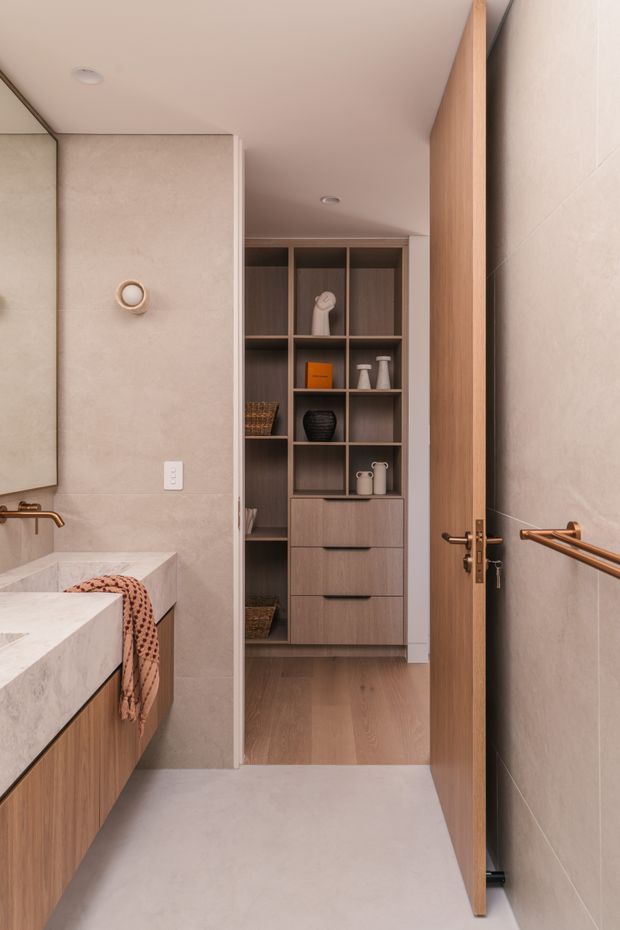 A refined contemporary bathroom viewed toward an open timber door and adjoining built-in storage. The bathroom features warm beige stone-look wall and floor finishes, a floating timber vanity with a pale stone benchtop, brushed bronze tapware and towel rails, and a full-height dark timber shelving and drawer unit beyond. Soft recessed lighting and understated decorative objects create a calm, warm, minimalist atmosphere.