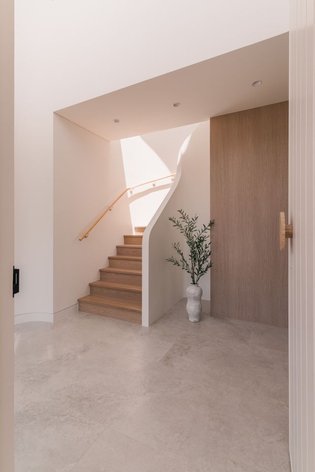 A refined minimalist residential stair hall viewed from an entry or circulation space. Warm oak-toned timber stairs rise beside a sculptural curved white balustrade, with a slim timber handrail and recessed ceiling downlights. Pale plaster walls and a large vertical timber-panelled wall create a calm, warm-neutral interior. A simple ceramic vase with greenery sits at the base of the stairs.
