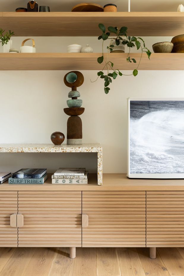 Close-up interior view of a warm, contemporary timber entertainment and storage unit. Horizontal wood shelving displays ceramics, books, decorative objects, and a trailing plant above a fluted timber credenza with slim legs. A terrazzo-edged open niche and framed ocean artwork add texture and visual interest. The palette is natural and muted, with light timber, warm off-white walls, and earthy decorative accents.