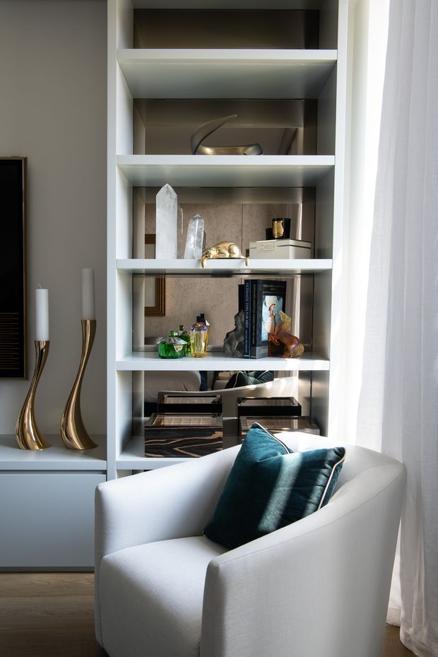 A refined living room vignette featuring a white upholstered armchair with a deep teal cushion positioned in front of illuminated built-in open shelving. The shelving displays books, sculptural decor, crystals, metallic objects, and decorative boxes, with a nearby console and tall gold-toned candleholders. Natural light filters through sheer white curtains, creating a calm, luxurious atmosphere.