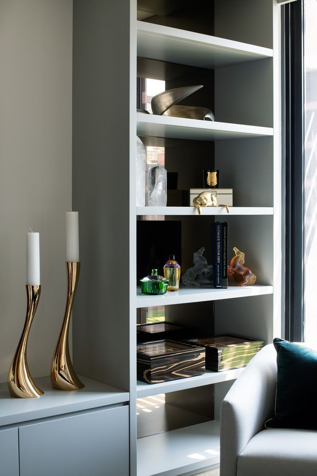 A close-up interior detail of a contemporary built-in shelving unit beside a large window. The open white shelving displays sculptural objects, books, glassware and decorative pieces, while integrated lower cabinetry provides concealed storage. Two tall metallic candleholders sit on the adjacent built-in cabinet, and part of a light upholstered armchair with a dark cushion is visible in the foreground. Strong natural light creates a refined, curated atmosphere.
