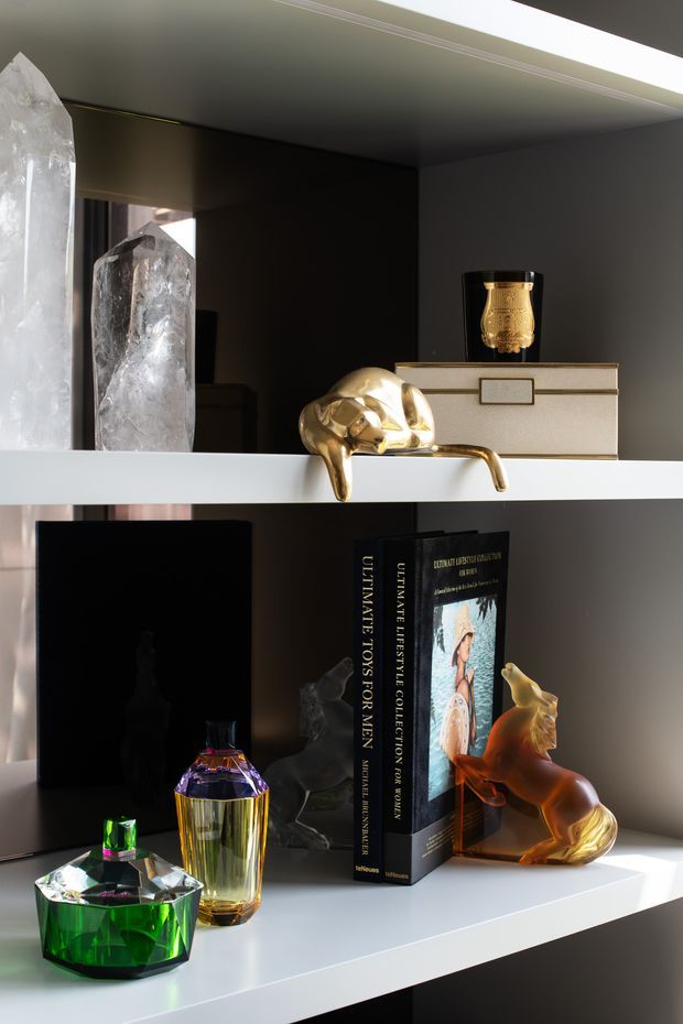Close-up interior detail of built-in white shelving displaying decorative crystal formations, books, perfume bottles, sculptural objects, and a gold-toned animal figurine. The shelves are set against a dark wall and softly illuminated, creating a refined, dramatic display vignette.