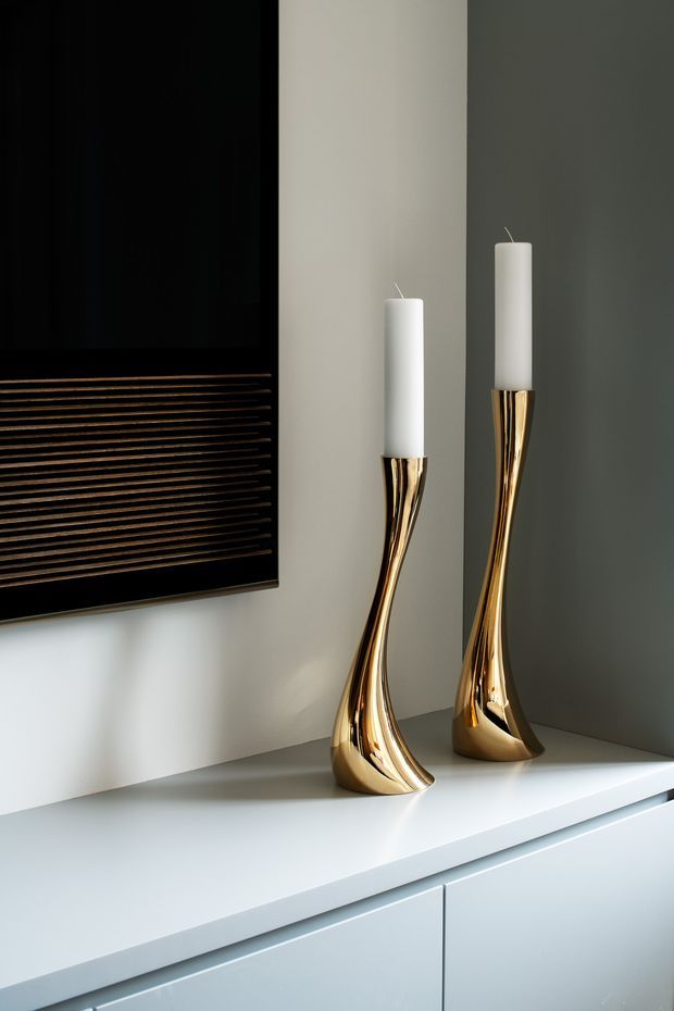 Close-up interior detail of two tall sculptural gold candleholders with white taper candles displayed on a white built-in console. A dark wall-mounted screen or panel with horizontal detailing is partially visible at left, with a reflective dark wall surface behind the candleholders. The composition is refined and minimalist with strong metallic accents.