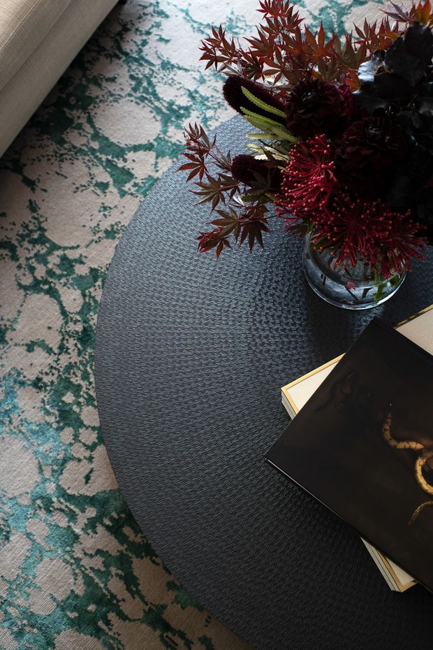 A close-up styled interior vignette showing a dark oval coffee table with a textured surface, a glass vase of deep red and burgundy foliage, and stacked books. A patterned rug in cream and teal sits beneath the table, with the edge of a light upholstered sofa visible at the upper left. The composition is moody and refined, emphasizing layered texture and rich contrasting colour.