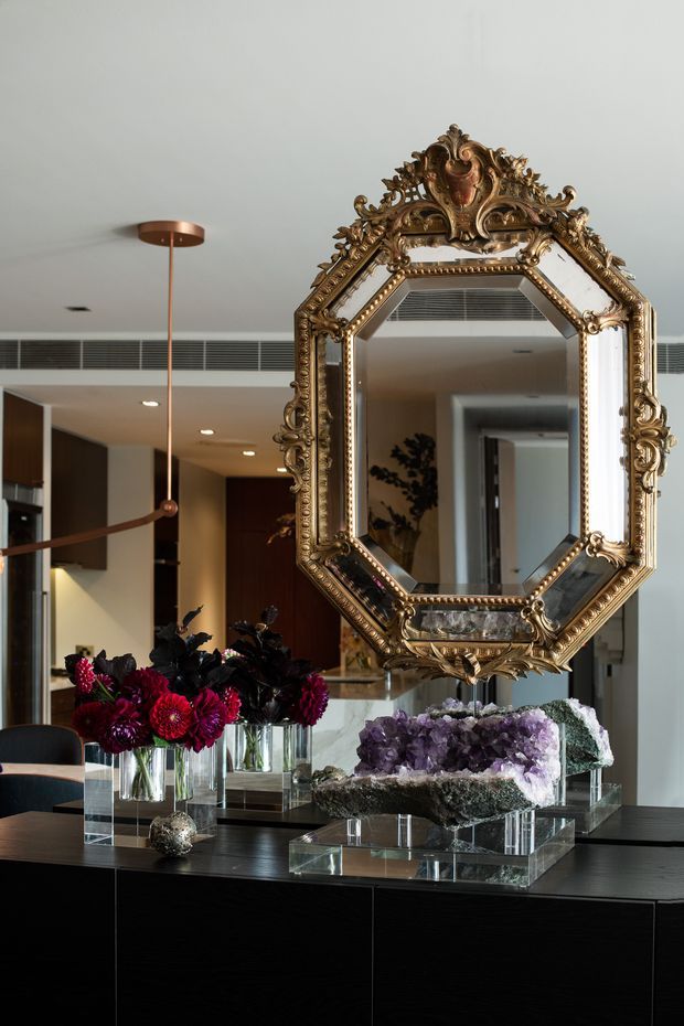 A sophisticated interior featuring a dark built-in console or sideboard beneath an ornate antique-style gold mirror. The console displays cut-flower arrangements in glass vases, large purple crystal-like decorative pieces on clear plinths, and a small metallic sculpture. A contemporary open-plan kitchen and hallway are visible in the mirror and background, with dark timber cabinetry, pale stone surfaces, recessed lighting, and a sculptural copper pendant light. The space combines richly decorative statement pieces with a refined contemporary interior.