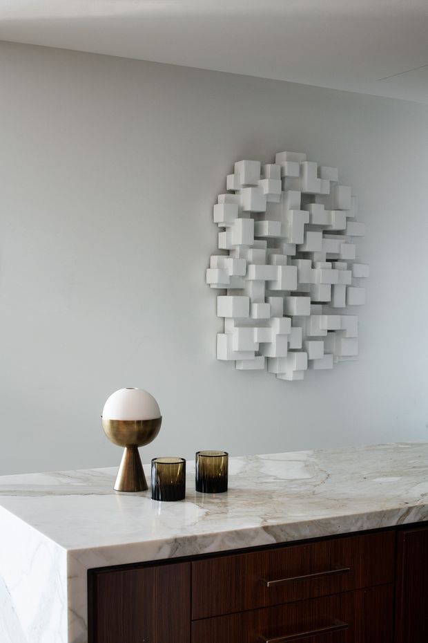 Close-up interior view of a refined built-in dark timber cabinet or sideboard with a thick white veined stone benchtop. A sculptural white three-dimensional wall installation is mounted above, accompanied by a brass-and-white table lamp and dark glass vessels. The composition is minimalist and luxurious, with a restrained neutral palette and warm wood accents.