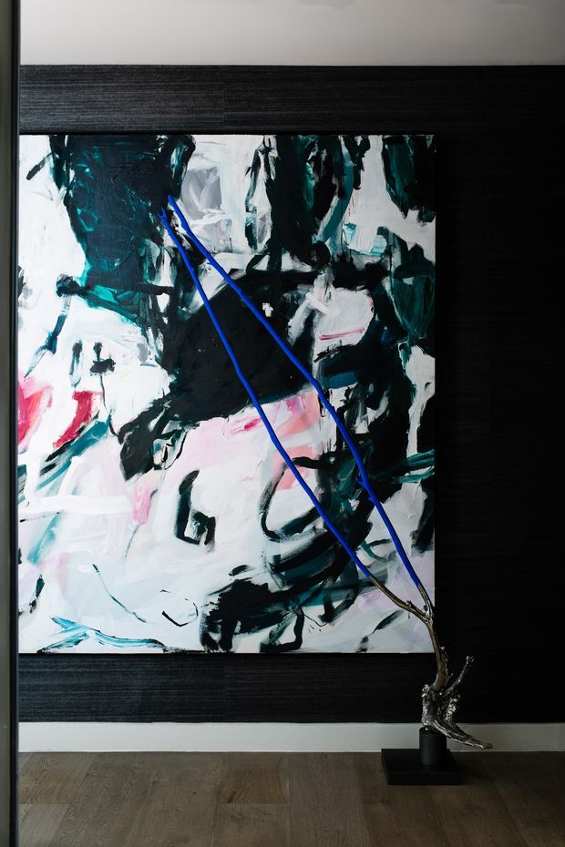 A dramatic interior feature wall with a large abstract artwork in expressive black, white, teal, pink, and blue brushstrokes. The artwork is framed by dark horizontal textured panels and a pale border, with a sculptural metallic floor piece positioned in front. Timber flooring and strong black-and-white contrast create a sophisticated, gallery-like atmosphere.