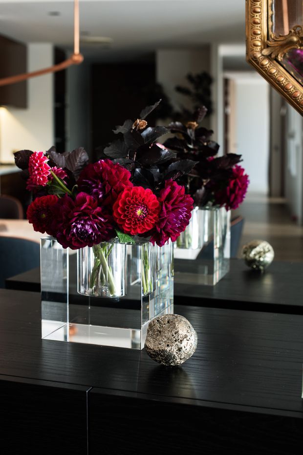 A sophisticated interior vignette featuring deep burgundy and magenta flowers arranged in a clear rectangular glass vase on a dark, glossy console or dining table. A large wall mirror reflects the arrangement and surrounding contemporary space, while an ornate gold decorative frame appears at the upper right and metallic spherical ornaments add contrast. The palette is dramatic and refined, with a modern interior accented by luxe decorative elements.