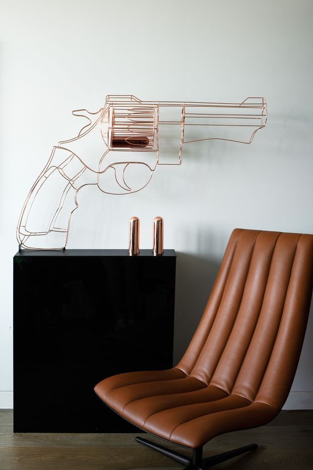 A stylised interior vignette featuring a sculptural brown leather lounge chair with deep vertical channel tufting, a glossy black cabinet, two copper-toned cylindrical objects, and a large minimalist linear revolver artwork mounted on a pale wall. Dark timber flooring and restrained furnishings create a dramatic, gallery-like composition.