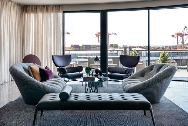 A refined contemporary living room with two sculptural curved sofas, matching lounge chairs, a tufted ottoman, and a glass coffee table arranged on a large rug. Full-height glazing overlooks an urban industrial waterfront or port setting, while sheer floor-to-ceiling curtains soften the room. The interior uses dark upholstered furniture, pale tiled flooring, and a restrained neutral palette.