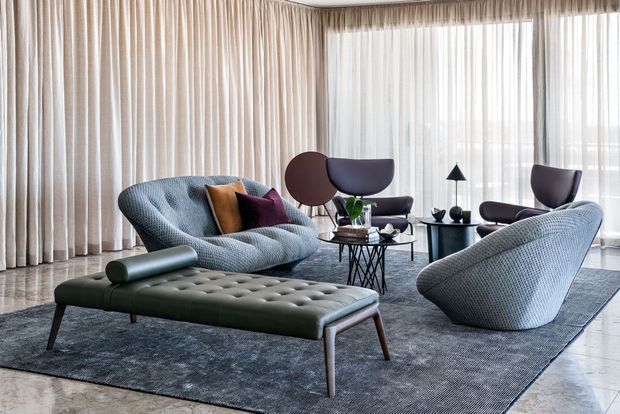 A refined living room arranged around a large tufted ottoman-style coffee table, with sculptural upholstered lounge chairs, a small round coffee table, and dark side tables. Full-height sheer and draped curtains cover expansive windows, while a large grey rug anchors the seating area over a polished light floor. The palette is muted and sophisticated, with blue-grey upholstery, charcoal grey textiles, and warm neutral curtains.