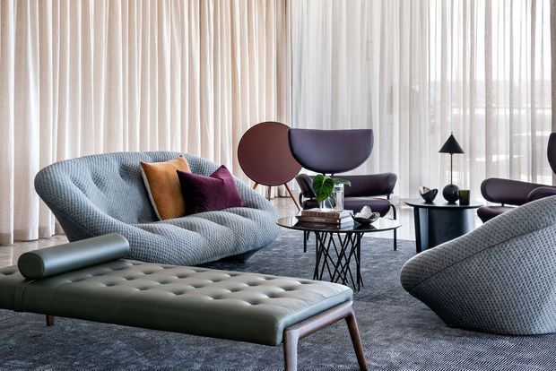 A refined mid-century-inspired living room furnished with sculptural lounge chairs, a curved textured sofa, a tufted leather ottoman, nesting coffee tables, and a minimalist floor lamp. Full-height sheer curtains diffuse abundant daylight across a grey carpet and muted neutral palette, with burgundy and mustard cushions providing accents.
