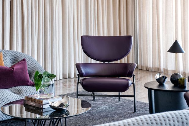 Refined contemporary living room with a sculptural dark plum lounge chair, curved textured sofa, round glass coffee table, black side table, and minimalist floor lamp. Full-height sheer curtains create a softly lit backdrop, while purple upholstery and dark accents contrast with pale neutral furnishings and flooring.