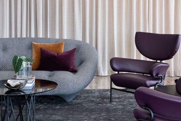 A refined living room vignette featuring a sculptural blue-grey curved sofa, deep plum leather lounge chairs, and layered plum and ochre cushions. Floor-to-ceiling cream curtains, a dark textured rug, glass-topped black coffee tables, and a small indoor plant create a sophisticated, moody seating area.