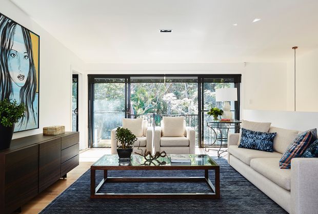 Bright contemporary living room with a large cream sectional sofa, two upholstered armchairs, a dark rectangular glass-top coffee table, and a deep charcoal area rug. Floor-to-ceiling black-framed glazing opens to leafy greenery outside, while a dark timber sideboard, oversized portrait artwork, indoor plant, and floor lamp complete the refined interior.