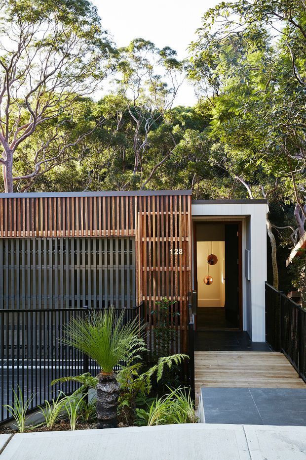 Street-facing view of a contemporary residential entrance integrated into a leafy bush setting. The facade combines vertical timber slats, dark metal screening, white rendered or painted wall surfaces, and a recessed entry door reached by a timber and dark tiled walkway. Layered planting softens the entrance, while the number 128 and warm pendant lights are visible inside the doorway.