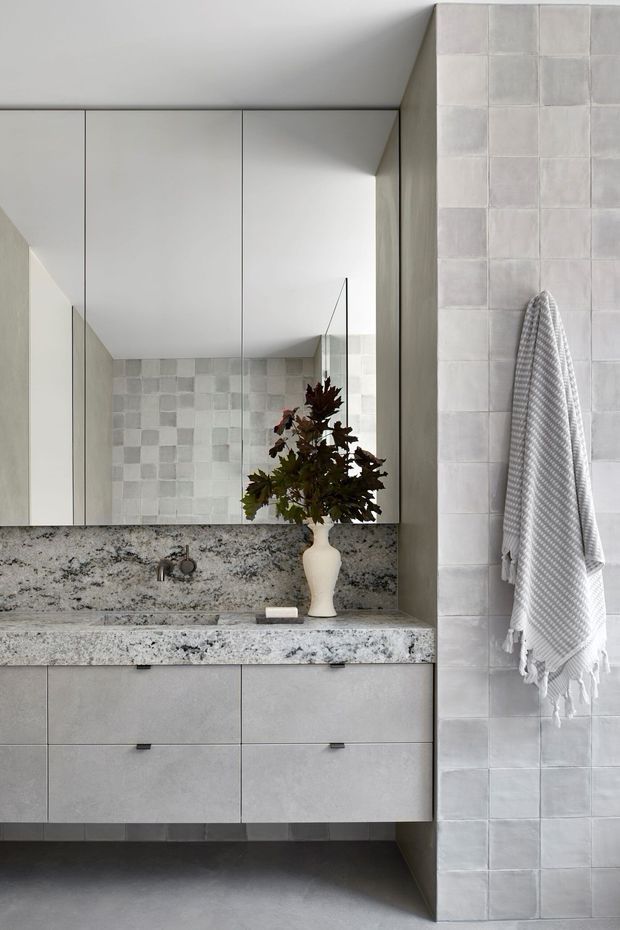 A contemporary bathroom vanity with a large frameless mirror, floating grey cabinetry, and a heavily patterned pale stone benchtop and integrated basin. The walls feature pale grey square tiles with subtle tonal variation, while a textured white towel hangs on the tiled wall. A small decorative vase with foliage sits on the vanity, adding an organic accent to the otherwise restrained, monochromatic space.