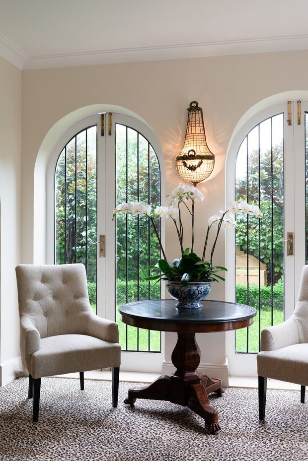 Elegant sitting area with two upholstered armchairs flanking a dark wood pedestal table, positioned before tall arched glazed doors with black metal grilles. The room features warm beige walls, white architectural trim, a decorative wall-mounted lantern, and a patterned carpet, creating a refined traditional interior.