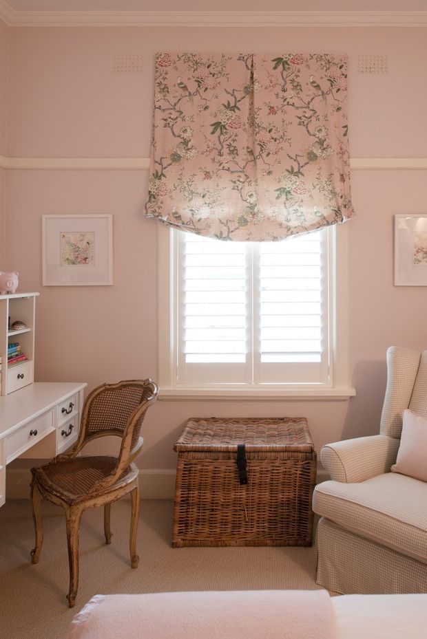 A charming pink-toned traditional sitting or reading room with a floral Roman blind over a white plantation-shuttered window. The room includes a white built-in desk and shelving, a woven rattan chair, a wicker storage trunk, and a slipcovered upholstered armchair. Decorative framed botanical artwork, wall moulding, carpeted flooring, and soft warm lighting create a cosy, refined atmosphere.