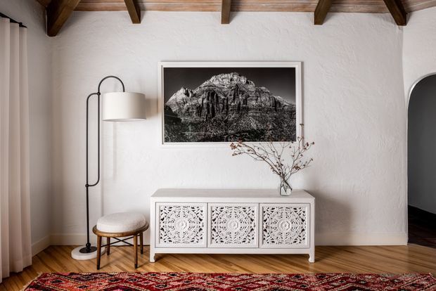 A warmly styled living room or lounge area with white textured plaster walls, exposed dark timber ceiling beams, honey-toned timber flooring, and a white carved-front sideboard beneath a framed black-and-white mountain photograph. A tall black floor lamp, upholstered stool, vase with branches, curtains, and a patterned red rug complete the space. A dark opening at right suggests an adjoining room or hallway.