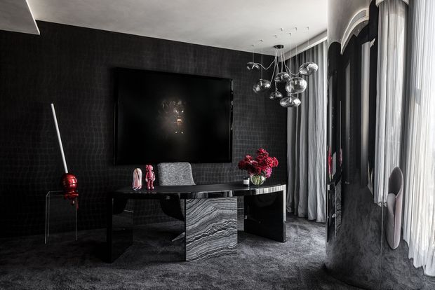A dramatic, luxurious living room or media room with a large wall-mounted television, black textured feature walls, dark carpet, a sculptural black console with a marbled stone central panel, and clustered chrome pendant lights. Floor-to-ceiling sheer curtains and a tall arched window or glazed opening line one side of the room, while sculptural furnishings and red floral accents add contrast.