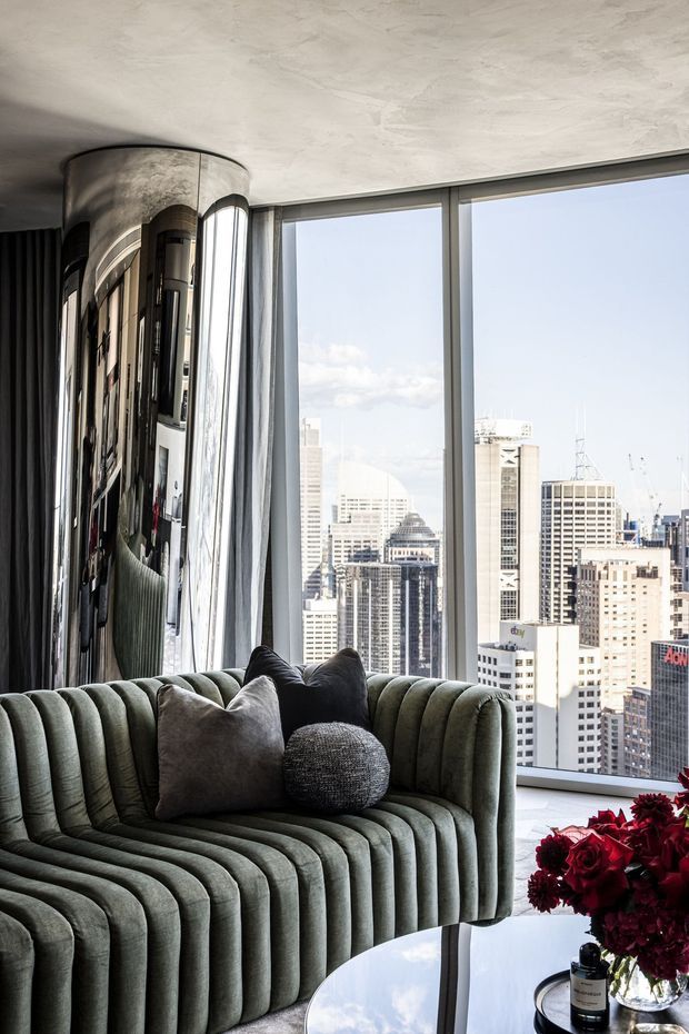 A sophisticated high-rise living room with a deeply channel-tufted sage-grey sofa, sculptural cushions, a round reflective coffee table, and dark red floral accents. Floor-to-ceiling windows provide expansive views of a dense urban skyline. A curved mirrored feature wall and textured plaster ceiling add a dramatic, luxurious character.