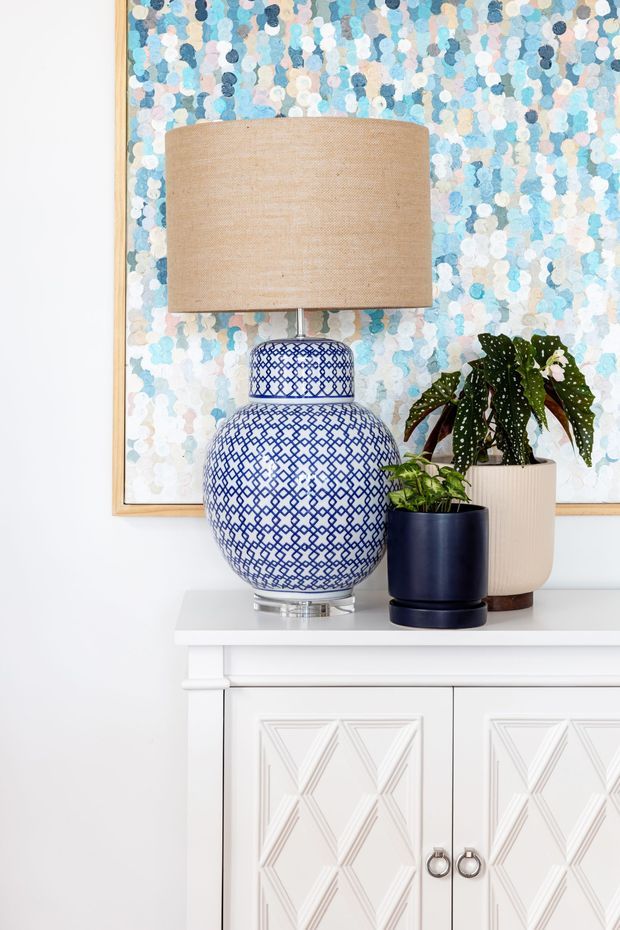 A bright styled interior vignette featuring a white decorative cabinet with geometric raised-panel doors, topped with a blue-and-white patterned ceramic lamp, navy and cream planters, and leafy indoor plants. A framed abstract artwork in soft blue, white, and pale pink tones forms the backdrop.