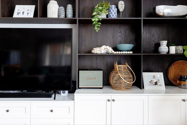 Built-in living room media and display cabinetry featuring a large television, dark timber open shelving, and white lower cabinets with drawers and doors. The shelves contain curated decorative objects, framed photographs, ceramics, a woven basket, and a compact speaker, creating a high-contrast, refined contemporary interior.
