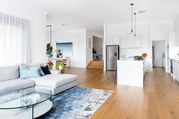 Bright open-plan living, dining, and kitchen interior with white cabinetry, a central island, stainless-steel appliances, timber flooring, high ceilings, and abundant sheer-curtained daylight. A pale sectional sofa and patterned blue rug define the living area, while simple pendant lights and classic trim add a refined contemporary feel.