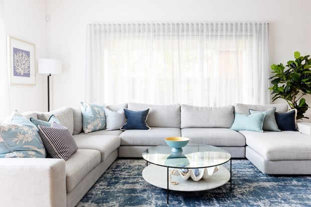 Bright, airy living room featuring a large light-grey L-shaped sectional sofa, blue and pale patterned cushions, a round glass coffee table with a sculptural white base, and a blue patterned rug. Sheer floor-to-ceiling curtains cover a large window, with a tall indoor plant, floor lamp, and framed artwork completing the space.