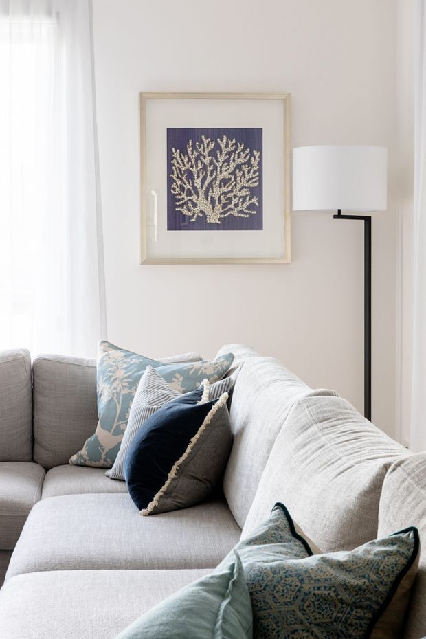 A bright, softly styled living room corner featuring a large light-grey sectional sofa with blue, teal, and patterned cushions. White sheer curtains frame a window, while a slim black floor lamp and framed coral artwork add simple decorative accents against pale neutral walls.
